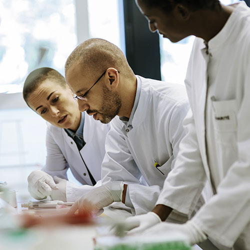 3 people in white lab coats and white gloves looking at something on the table